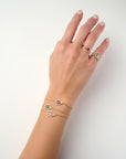 Hand adorned with gold rings, including a ruby-accented geometric design and a diamond-studded band, paired with three delicate gold chain bracelets featuring ruby, emerald, and diamond-shaped charms